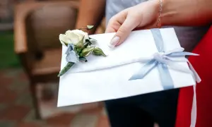 Dłoń trzyma białą kopertę z bukiecikiem i błękitną wstążką. Zastanawiasz się, ile chrzestny daje na wesele?