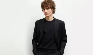 Młody mężczyzna w czarnym garniturze i czarnej koszuli, z rękami w kieszeniach, prezentuje elegancki styl.