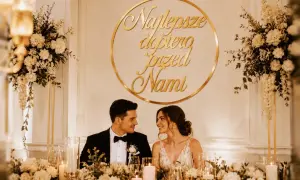 Para młoda, świeżo po ślubie, uśmiecha się do siebie przy udekorowanym stole. Złoty napis "Najlepsze dopiero przed nami" podkreśla ich szczęście.