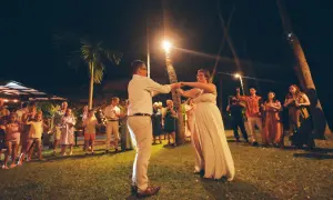 Para taniec nowożeńców podczas ślubu w plenerze. Goście obserwują parę na tle palm i nocnego nieba.
