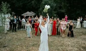 Panna młoda w bieli rzuca bukiet, a goście w barwnych strojach czekają na jego złapanie.