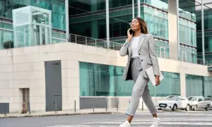 Kobieta w eleganckim stroju business casual rozmawia przez telefon, niosąc laptopa. Idzie ulicą przed nowoczesnym budynkiem.