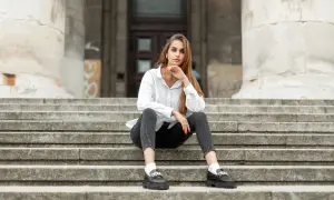 Eleganckie mokasyny to idealne buty do szarych spodni. Kobieta w białej koszuli siedzi na schodach.