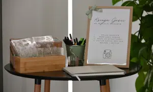 Stolik z księgą gości, pisakami i ozdobami na wesele. Na ramce instrukcja: "Wsuń obiektyw, zrób sobie zdjęcie, wklej do księgi gości, zostaw krótki wpis dla Michasi i Krzysia".