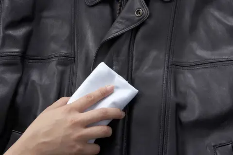 Dłoń z białym materiałem delikatnie naciera czarną skórzaną kurtkę, pokazując domowe sposoby, czym natłuścić kurtkę skórzaną.