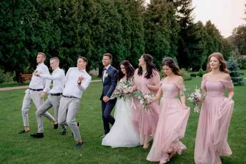 Para młoda i drużyna weselna w eleganckim ubiorze na ślub, idący przez trawnik.