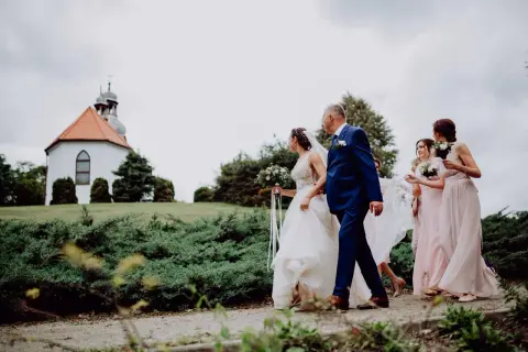 Panna młoda z bukietem idzie z ojcem do kościółka. Obok niej idzie świadkowa i druhny w różowych sukniach.