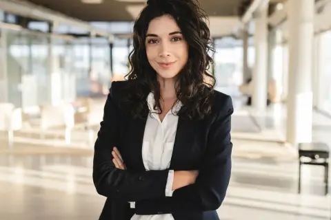 Kobieta w eleganckim business attire, z rękami skrzyżowanymi na piersi, pewnie patrzy w obiektyw.