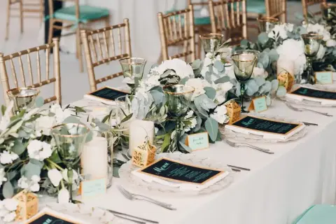Elegancki bankiet co to? Stół nakryty białym obrusem, złote krzesła, zielone kieliszki, menu i kwiaty.