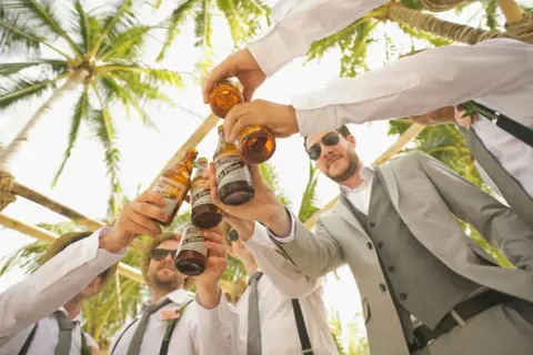 Mężczyźni w garniturach wznoszą toast butelkami piwa pod palmami. Idealny pomysł na kawalerski!
