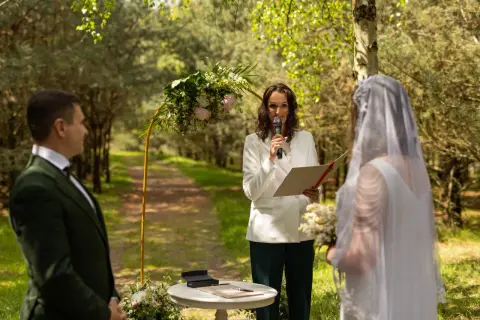 Uroczysta ceremonia ślubna w lesie. Pan młody w zielonym garniturze, panna młoda w białej sukni i welonie, a prowadząca ceremonię z mikrofonem i dokumentami.
