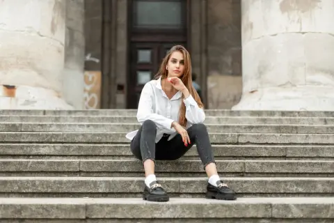 Eleganckie mokasyny to idealne buty do szarych spodni. Kobieta w białej koszuli siedzi na schodach.