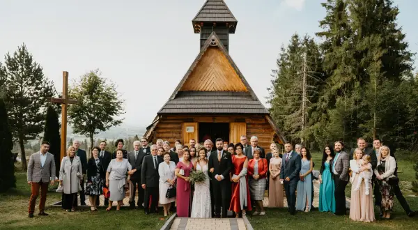 Uroczystości weselne przed drewnianą kapliczką. Para młoda w otoczeniu rodziny i przyjaciół.