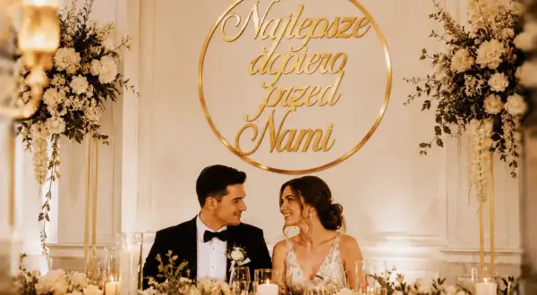 Para młoda, świeżo po ślubie, uśmiecha się do siebie przy udekorowanym stole. Złoty napis "Najlepsze dopiero przed nami" podkreśla ich szczęście.