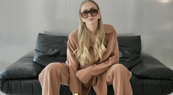 Elegancki biznesowy look damski: blondynka w beżowym garniturze, okularach i złotej biżuterii siedzi na czarnej kanapie.