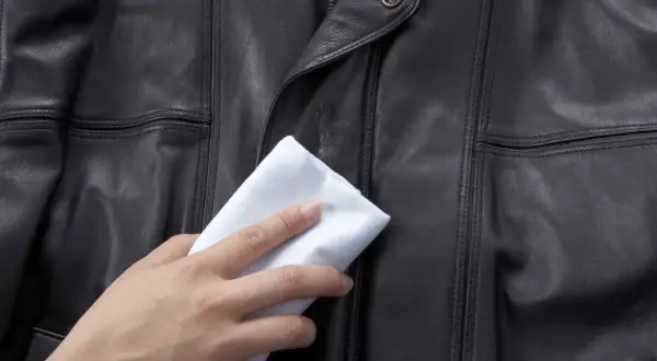 Dłoń z białym materiałem delikatnie naciera czarną skórzaną kurtkę, pokazując domowe sposoby, czym natłuścić kurtkę skórzaną.