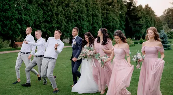 Para młoda i drużyna weselna w eleganckim ubiorze na ślub, idący przez trawnik.