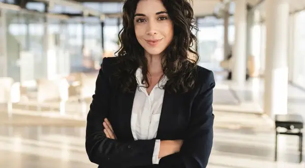 Kobieta w eleganckim business attire, z rękami skrzyżowanymi na piersi, pewnie patrzy w obiektyw.