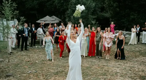 Panna młoda w bieli rzuca bukiet, a goście w barwnych strojach czekają na jego złapanie.