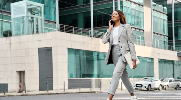 Kobieta w eleganckim stroju business casual rozmawia przez telefon, niosąc laptopa. Idzie ulicą przed nowoczesnym budynkiem.