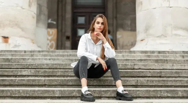 Eleganckie mokasyny to idealne buty do szarych spodni. Kobieta w białej koszuli siedzi na schodach.