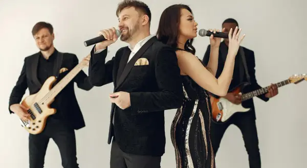 Zespół muzyczny w eleganckich strojach, wokalista i wokalistka śpiewają do mikrofonów, a gitarzyści grają na instrumentach. Idealni do oprawy muzycznej wesel i imprez, wykonają najlepsze przyśpiewki weselne.