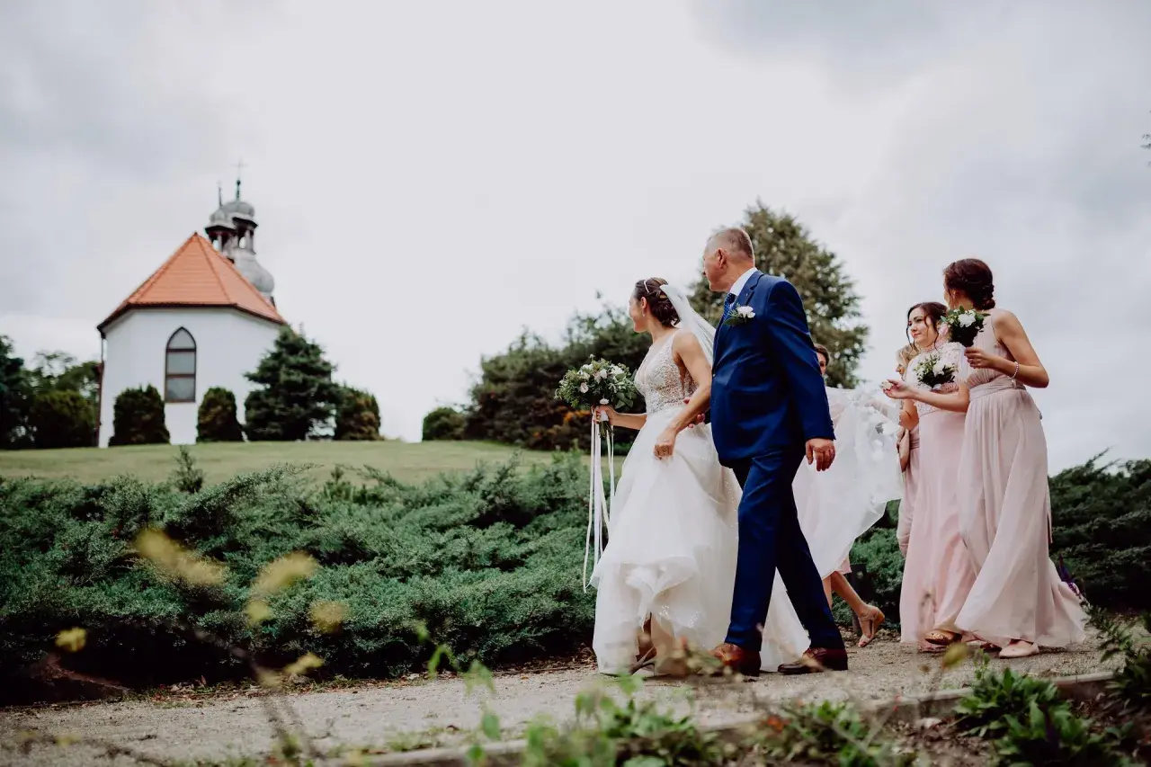 Panna młoda z bukietem idzie z ojcem do kościółka. Obok niej idzie świadkowa i druhny w różowych sukniach.