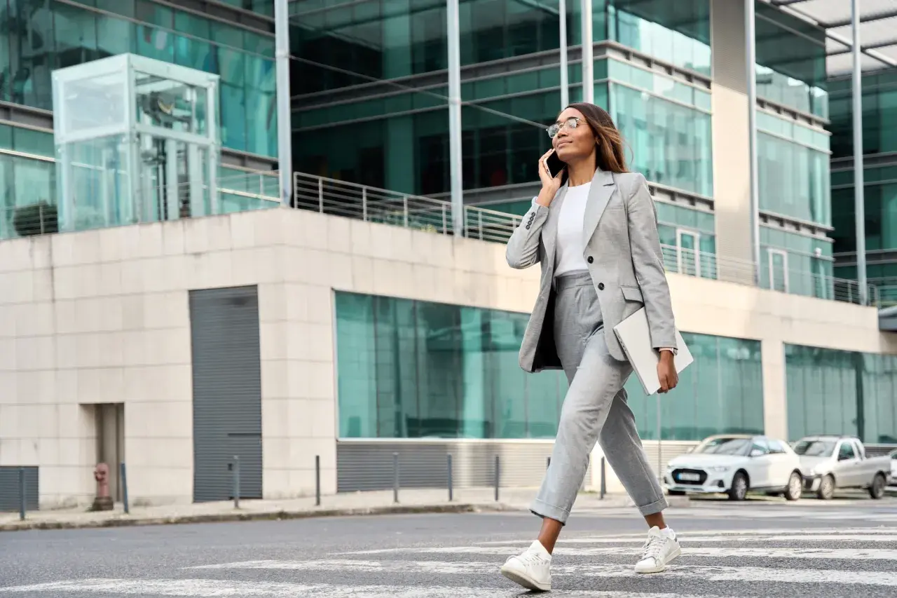 Kobieta w eleganckim stroju business casual rozmawia przez telefon, niosąc laptopa. Idzie ulicą przed nowoczesnym budynkiem.