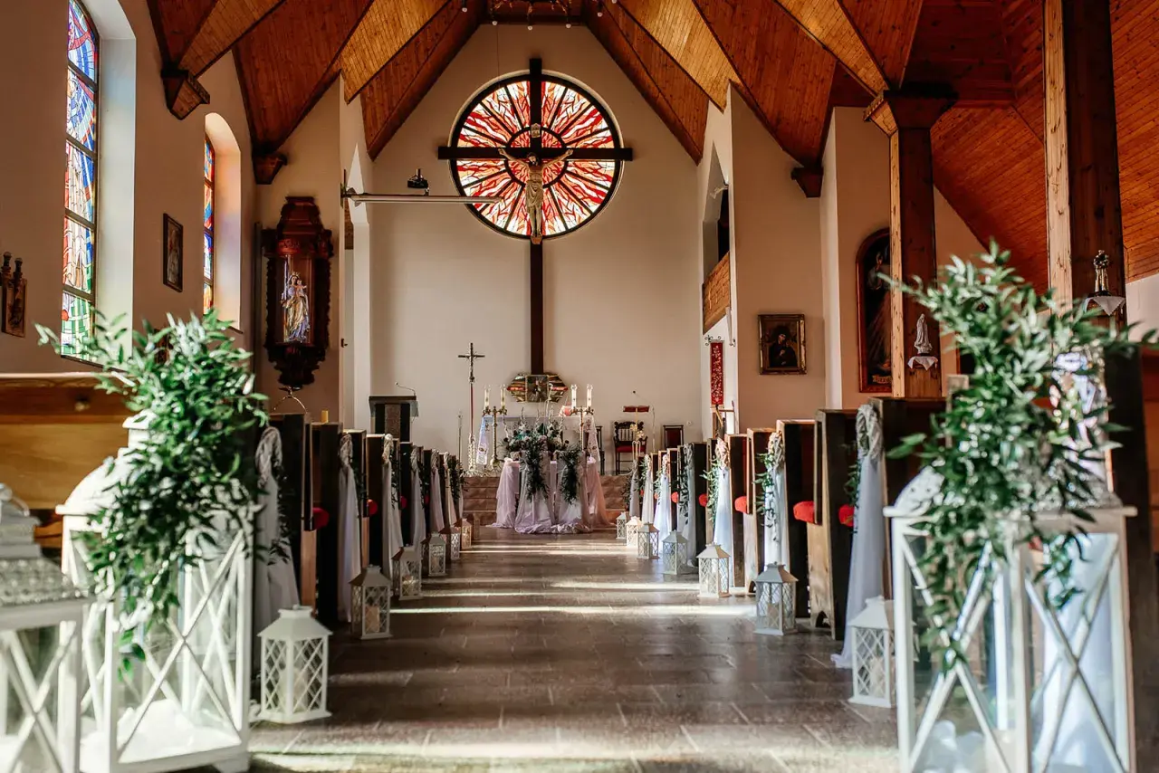 Elegancka dekoracja kościoła na ślub samemu: ławki ozdobione zielenią i lampionami, ołtarz z kwiatami.