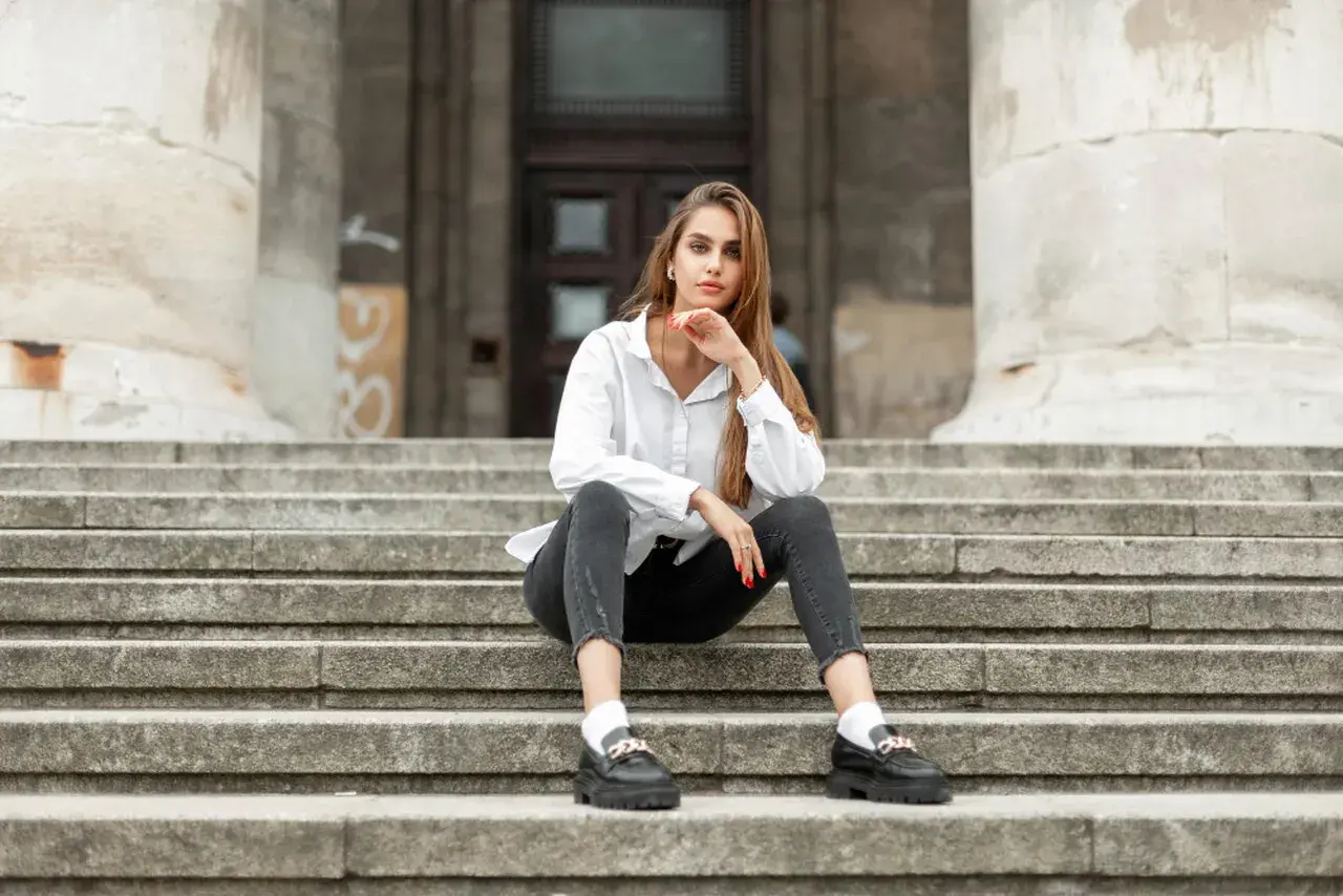 Eleganckie mokasyny to idealne buty do szarych spodni. Kobieta w białej koszuli siedzi na schodach.