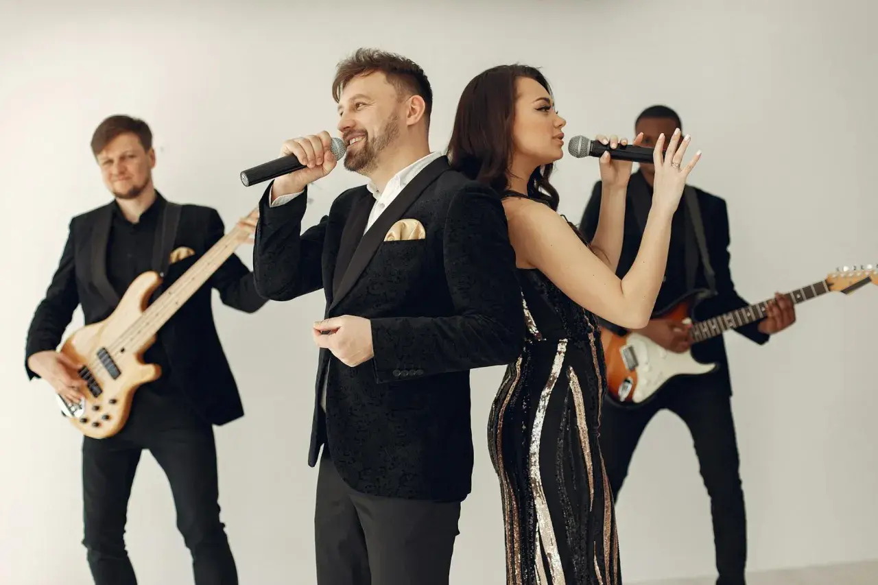 Zespół muzyczny w eleganckich strojach, wokalista i wokalistka śpiewają do mikrofonów, a gitarzyści grają na instrumentach. Idealni do oprawy muzycznej wesel i imprez, wykonają najlepsze przyśpiewki weselne.