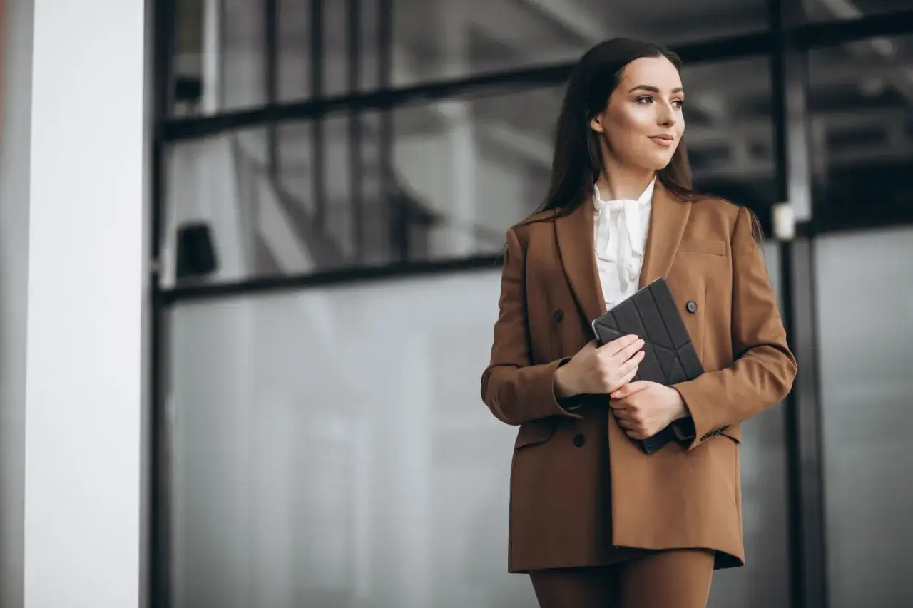 Elegancka kobieta w brązowej marynarce i białej bluzce, z tabletem w dłoniach. Jej biznesowy look emanuje pewnością siebie.