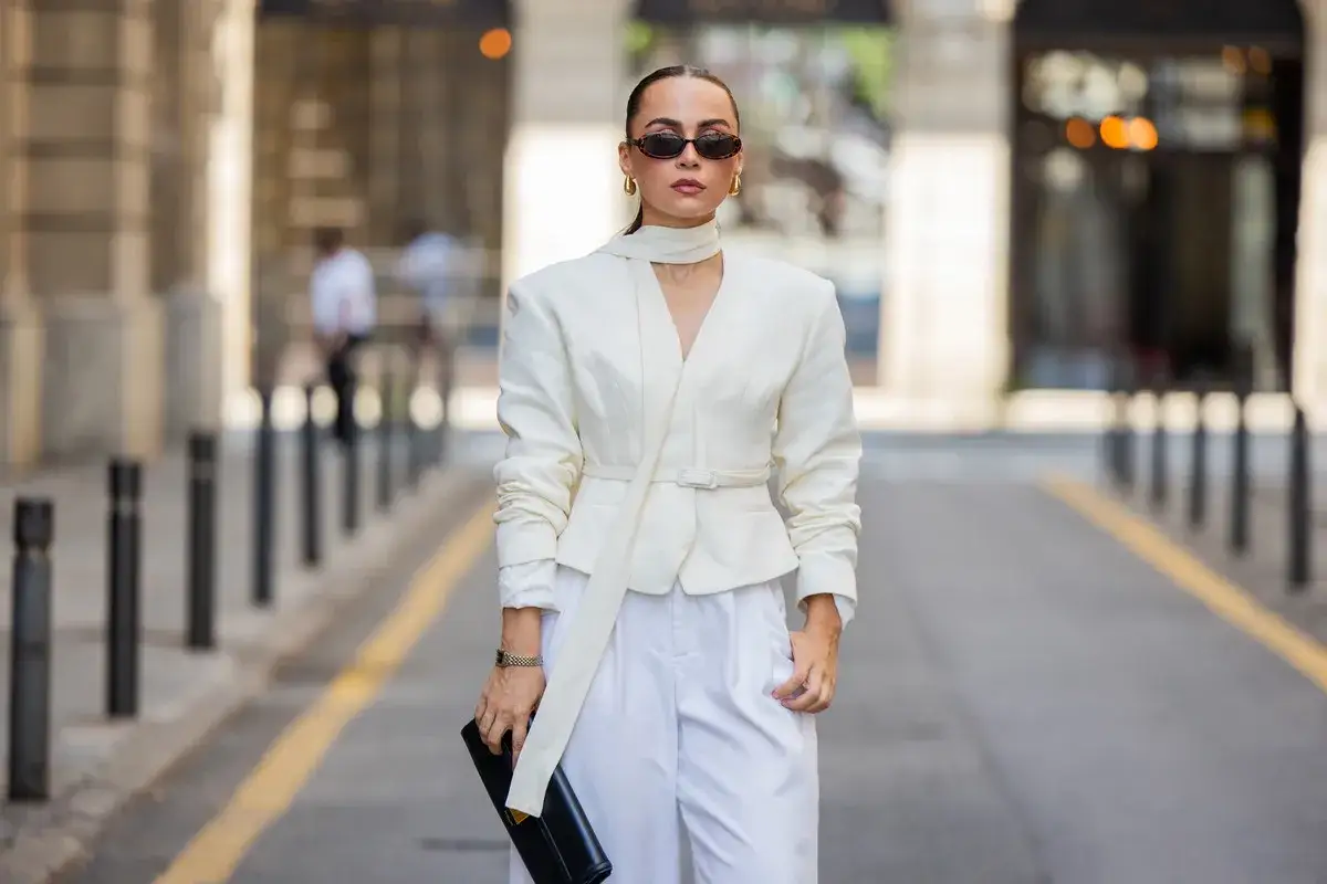 Modna it girl w białym, eleganckim komplecie, okularach i z czarną kopertówką na ulicy.