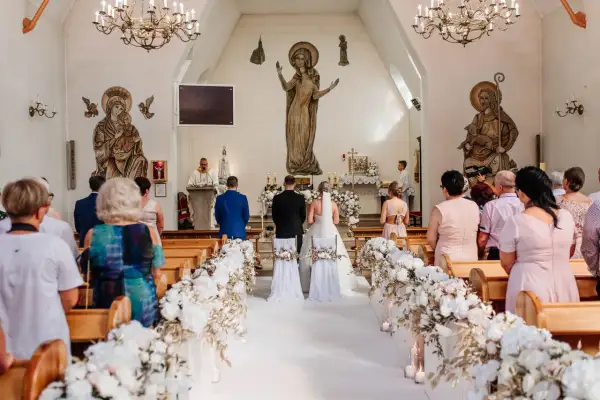 Przebieg mszy ślubnej: para młoda stoi przed ołtarzem w kościele, otoczona gośćmi i kwiatowymi dekoracjami.