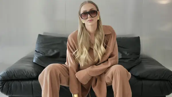 Elegancki biznesowy look damski: blondynka w beżowym garniturze, okularach i złotej biżuterii siedzi na czarnej kanapie.