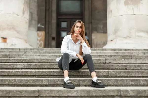 Eleganckie mokasyny to idealne buty do szarych spodni. Kobieta w białej koszuli siedzi na schodach.